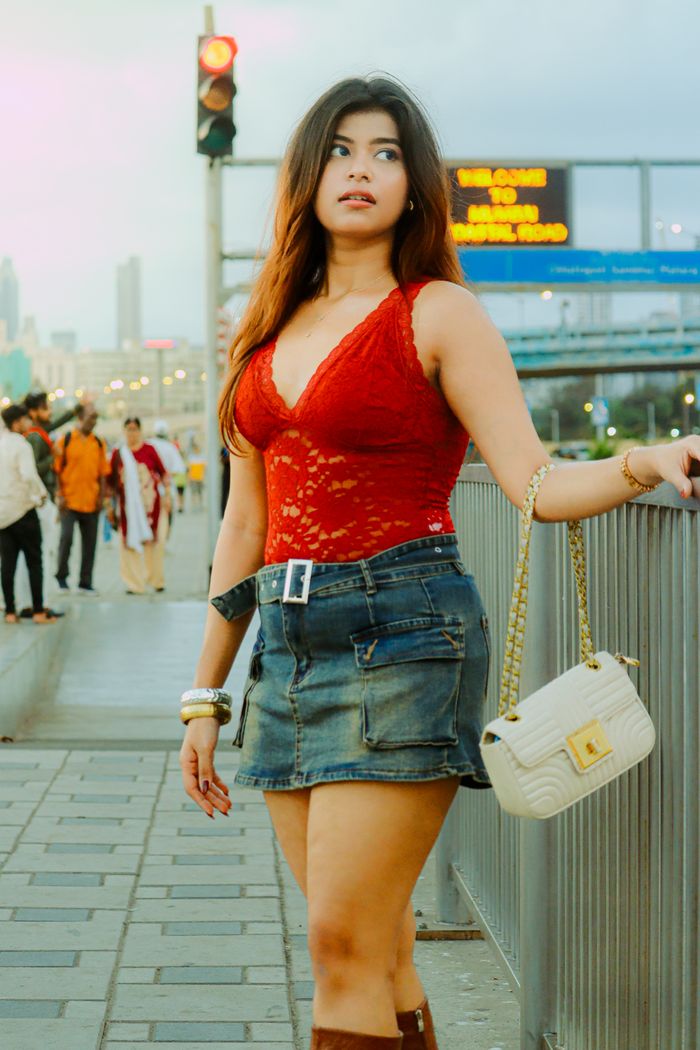 Stylish woman in vibrant red lace top and denim skirt walking on a bustling street in Mumbai, India.